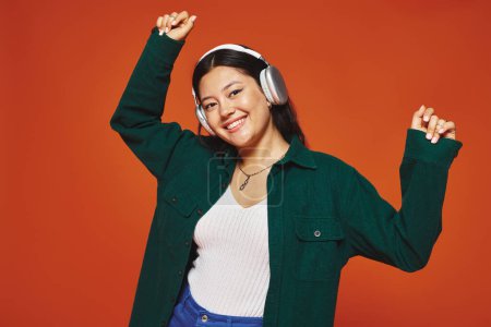 lustige und brünette asiatische Frau genießt Musik mit drahtlosen Kopfhörern auf orangefarbenem Hintergrund
