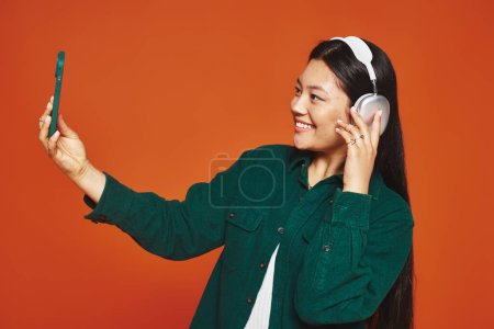 mujer asiática positiva disfrutando de la música con auriculares inalámbricos y tomando selfie sobre fondo naranja