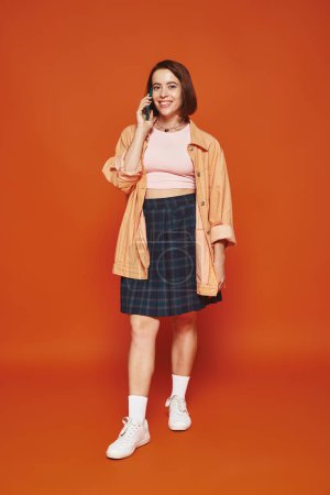 fröhliche junge Frau in stylischer Kleidung spricht auf Smartphone auf orangefarbenem Hintergrund, telefoniert