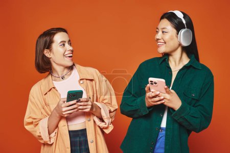 two happy multicultural friends using smartphones and listening music on orange background