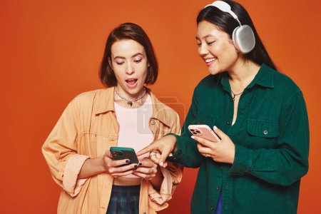 two cheerful multicultural friends using smartphones and listening music on orange background