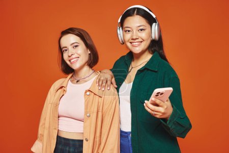 glückliche Asiatin mit drahtlosen Kopfhörern, die ihr Smartphone in der Nähe ihrer Freundin in Orange hält