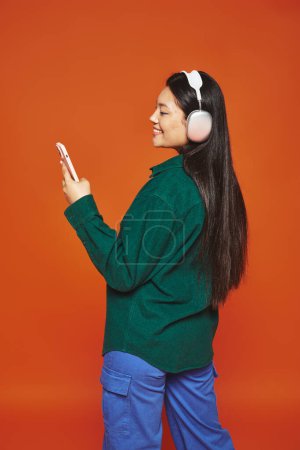 Téléchargez les photos : Heureux asiatique femme écoute de la musique dans sans fil casque en utilisant smartphone sur fond orange - en image libre de droit