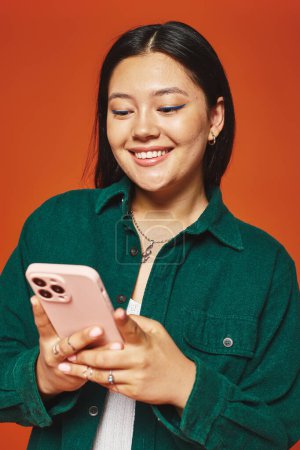 Téléchargez les photos : Heureux et jeune asiatique femme avec brune cheveux en utilisant smartphone et sourire sur fond orange - en image libre de droit