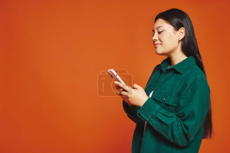 Téléchargez les photos : Joyeuse et jeune femme asiatique avec des cheveux bruns en utilisant un smartphone et souriant sur fond orange - en image libre de droit
