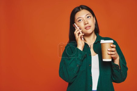 Téléchargez les photos : Jeune femme asiatique parler sur smartphone et tenant tasse en papier avec café sur fond orange - en image libre de droit