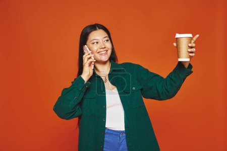 Téléchargez les photos : Heureux asiatique femme parler sur smartphone et tenant tasse en papier avec café sur fond orange - en image libre de droit