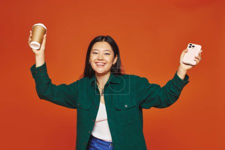Téléchargez les photos : Excité asiatique femme parler sur smartphone et tenant tasse en papier avec café sur fond orange - en image libre de droit