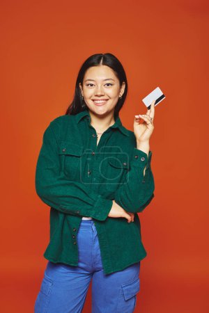 glückliche junge asiatische Frau in grünem Sakko mit Kreditkarte auf orangefarbenem Hintergrund, Konsumdenken