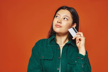 pensive young asian woman in green jacket holding credit card on orange background, consumerism