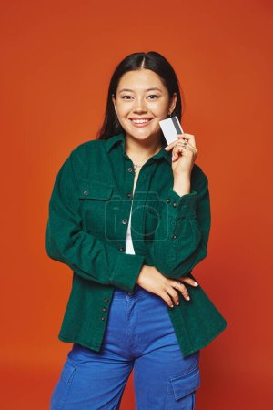 positive jeune femme asiatique en veste verte tenant carte de crédit sur fond orange, consumérisme