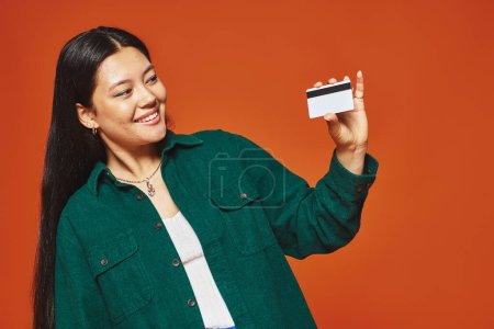 glückliche junge asiatische Frau in grünem Sakko mit Blick auf Kreditkarte auf orangefarbenem Hintergrund, Konsumdenken
