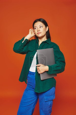 nachdenkliche junge asiatische Frau in lebendiger Kleidung mit Laptop auf orangefarbenem Hintergrund, Fernarbeit