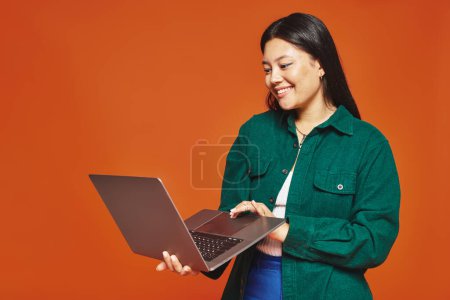 happy young asian woman in vibrant attire using laptop on orange background, remote work concept