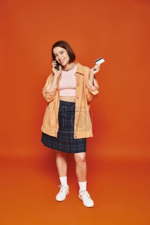 happy woman talking on smartphone and holding credit card on orange background, online shopping