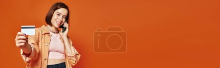 happy woman talking on smartphone and holding credit card on orange background, shopping banner