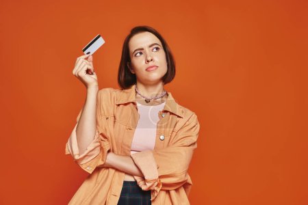 mujer joven pensativa con el pelo corto que sostiene la tarjeta de crédito en fondo naranja, finanzas personales