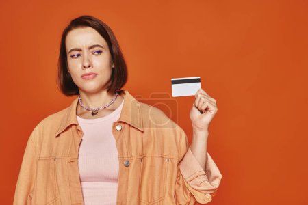 nachdenkliche junge Frau mit kurzen Haaren, Kreditkarte auf orangefarbenem Hintergrund, persönliche Finanzen