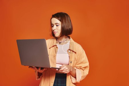 joven freelancer con atuendo casual sosteniendo laptop y trabajando remotamente sobre fondo naranja