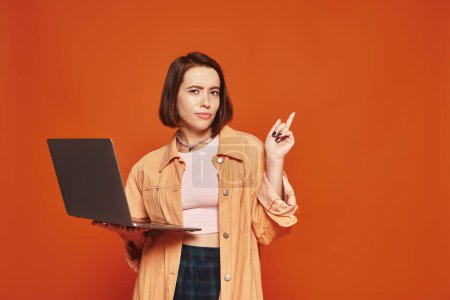 junge Freiberuflerin in Freizeitkleidung mit Laptop und Zeigefinger auf orangefarbenem Hintergrund