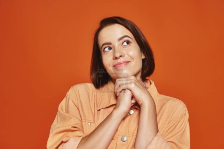 hopeful young woman with short hair smiling and looking away on orange background, cheerful