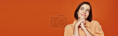 hopeful young woman with short hair smiling and looking away on orange background, banner