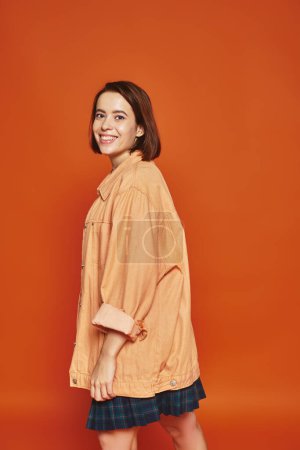 Foto de Mujer joven positiva con el pelo corto mirando a la cámara y sonriendo sobre fondo naranja, despreocupada - Imagen libre de derechos
