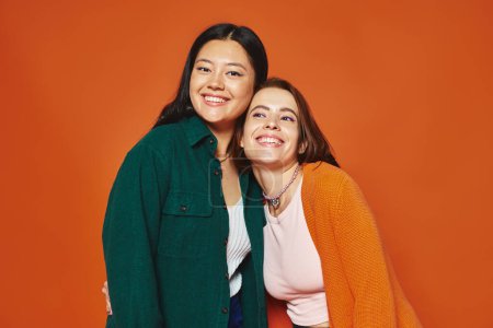radiant and multicultural female friends sharing a joyous moment on warm orange backdrop, diversity