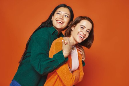 zwei Freundinnen in lässiger Kleidung, die sich umarmen und glückliche Momente miteinander auf orangefarbenem Hintergrund teilen