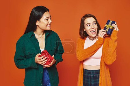 multicultural female friends exchanging gifts, smiling and having a good time on orange background