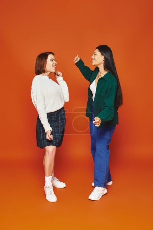 fröhliche multikulturelle Freundinnen in lebendiger Kleidung, die einander auf orangefarbenem Hintergrund anschauen