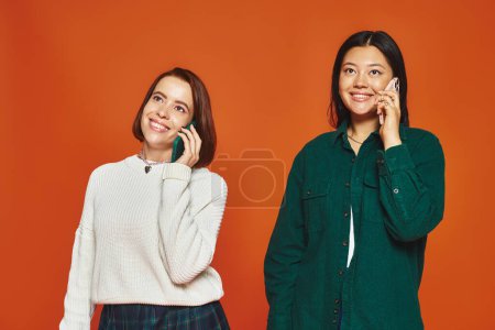 junge multikulturelle Frauen in lässiger und lebendiger Kleidung unterhalten sich auf Smartphone auf orangefarbenem Hintergrund