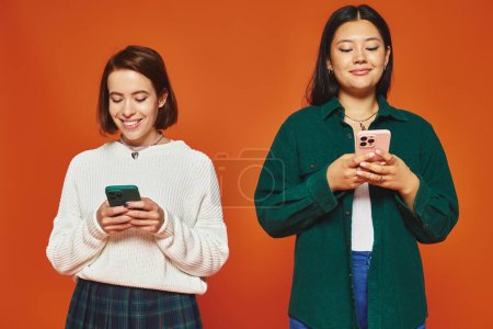 multicultural women using smartphones, engaged in social media on vibrant orange background