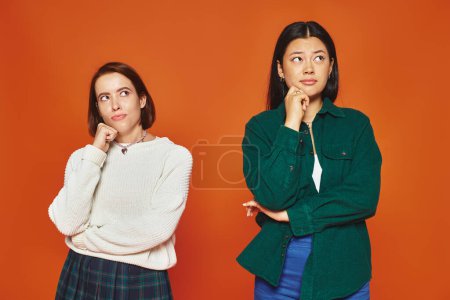 pensive young multicultural friends in vibrant attire looking away on orange background, think