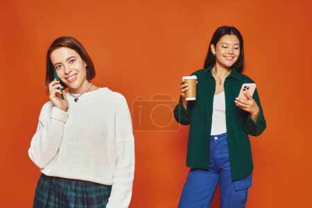 Foto de Mujeres alegres y jóvenes multiculturales utilizando teléfonos inteligentes y de pie sobre fondo naranja - Imagen libre de derechos