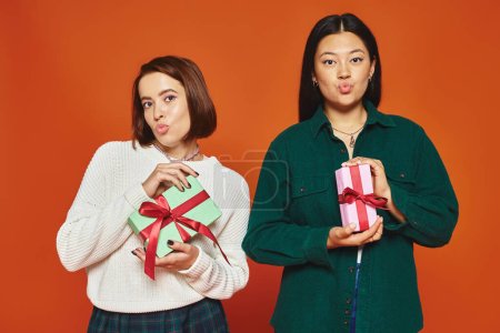 young multicultural female friends pouting lips and holding presents on orange background, festive