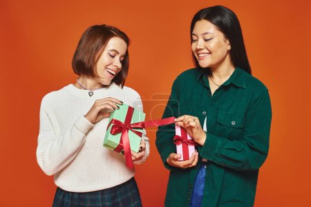 excited young multicultural female friends holding presents on orange background, gift exchange