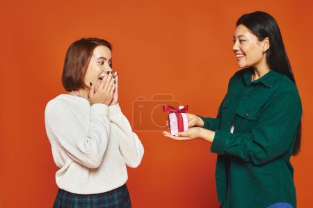 happy asian woman giving present to female friend covering face from surprise on orange background