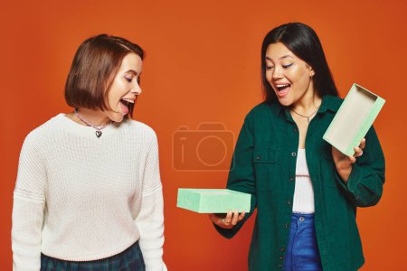 emocionado asiático mujer apertura presente cerca feliz mujer amigo en naranja fondo, regalo dando