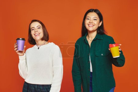 two multicultural female friends holding paper cups with coffee to go on orange background