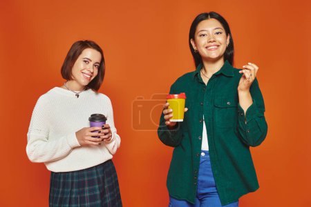 cheerful multicultural female friends holding paper cups with coffee to go on orange background