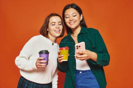 fröhliche multikulturelle Freundinnen, die Pappbecher mit Kaffee in der Hand halten und aufs Smartphone schauen