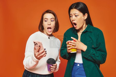 shocked multicultural female friends holding paper cups with coffee and looking at smartphone