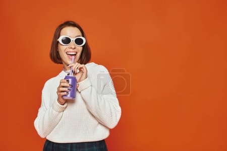 Stylish young woman in white trendy sunglasses sipping soda drink from can on orange backdrop