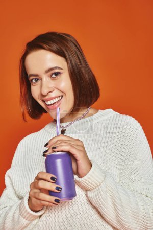 mujer joven con estilo en suéter de punto blanco sorbiendo bebida gaseosa de paja sobre fondo naranja