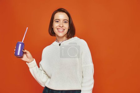 cheerful young woman in white knitted sweater holding soda can with straw on orange backdrop