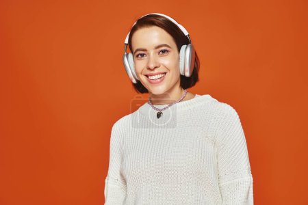cheerful young woman in white sweater and wireless headphones listening music on orange backdrop