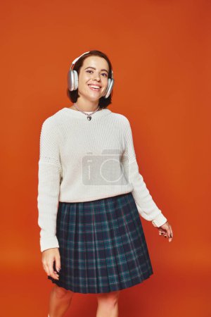 fröhliche junge Frau in weißem Pullover und kabellosen Kopfhörern, die Musik auf orangefarbenem Hintergrund hört