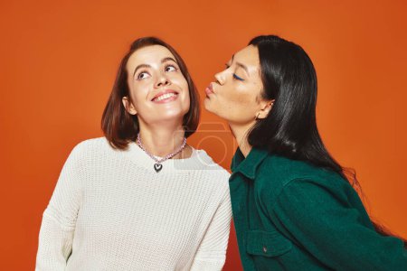 niedliche asiatische Frau küsst Wange ihrer fröhlichen Freundin auf orangefarbenem Hintergrund, Freundschaft