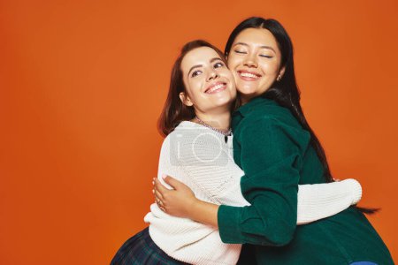 glückliche interrassische Freunde in lässiger Kleidung umarmen und lächeln auf orangefarbenem Hintergrund, Frauen